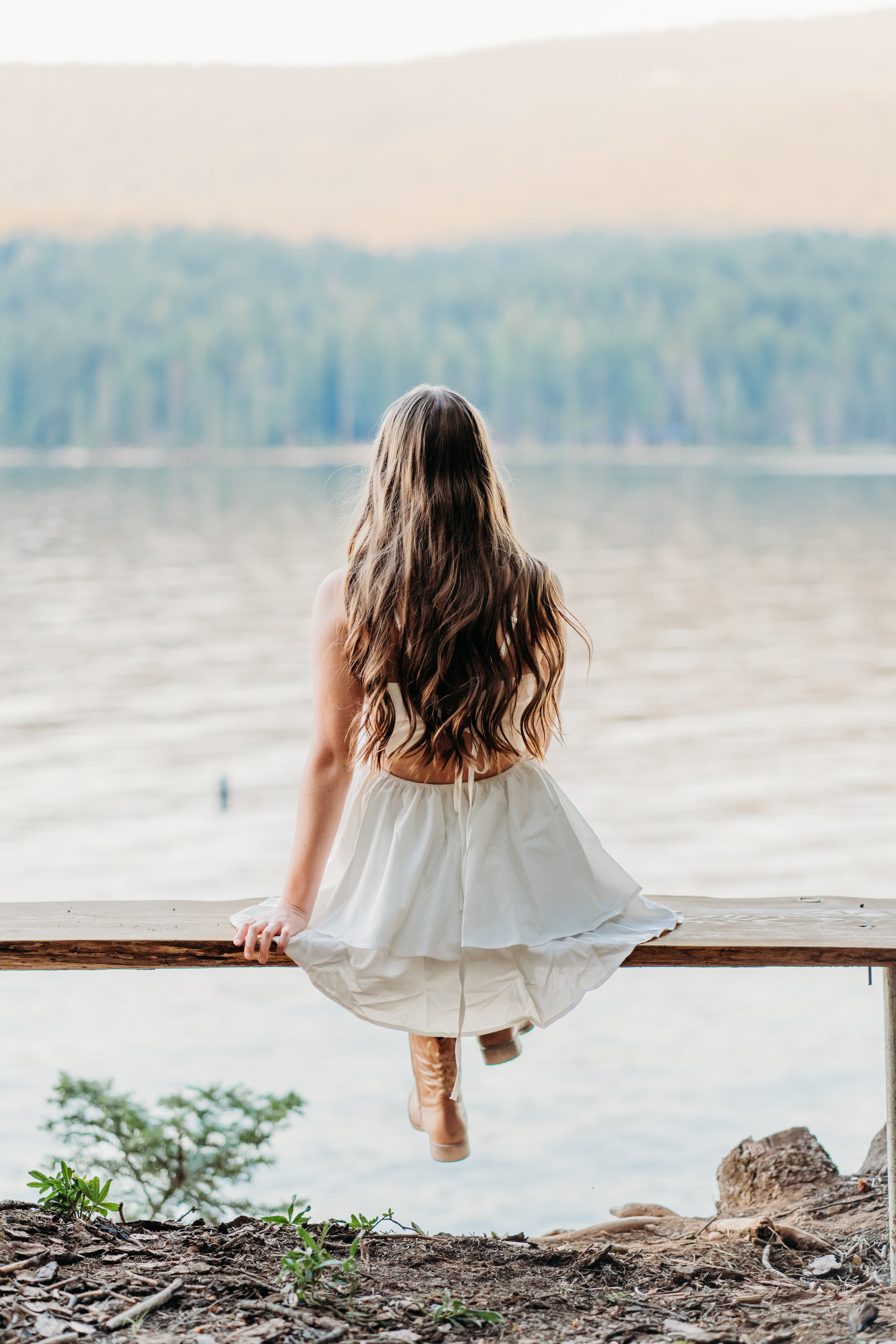 Young woman in a white dress sits on a dock rail over the lake, back to camera
