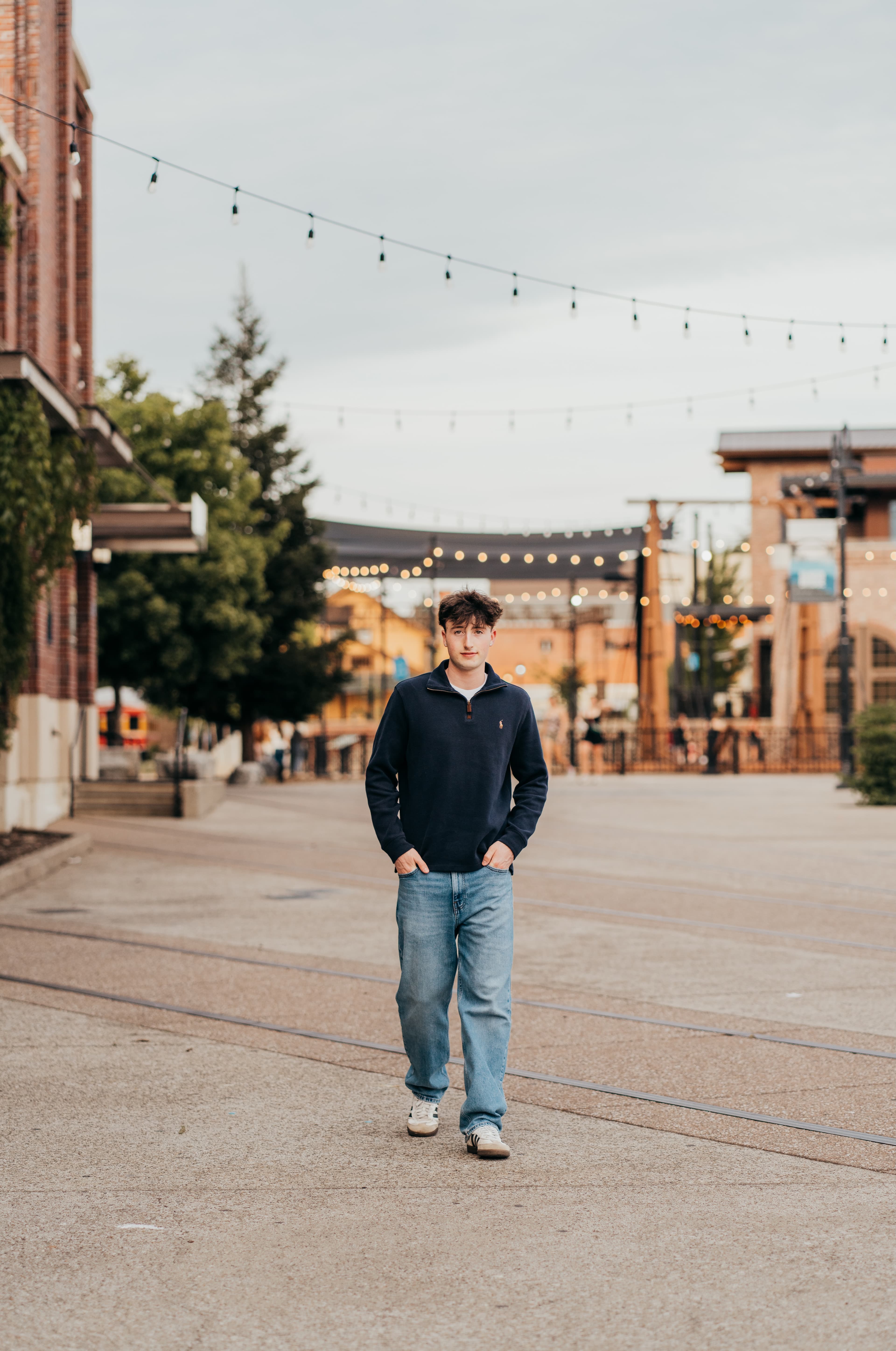 Young man in a navy quarter-zip walks through an urban plaza strung with string lights
