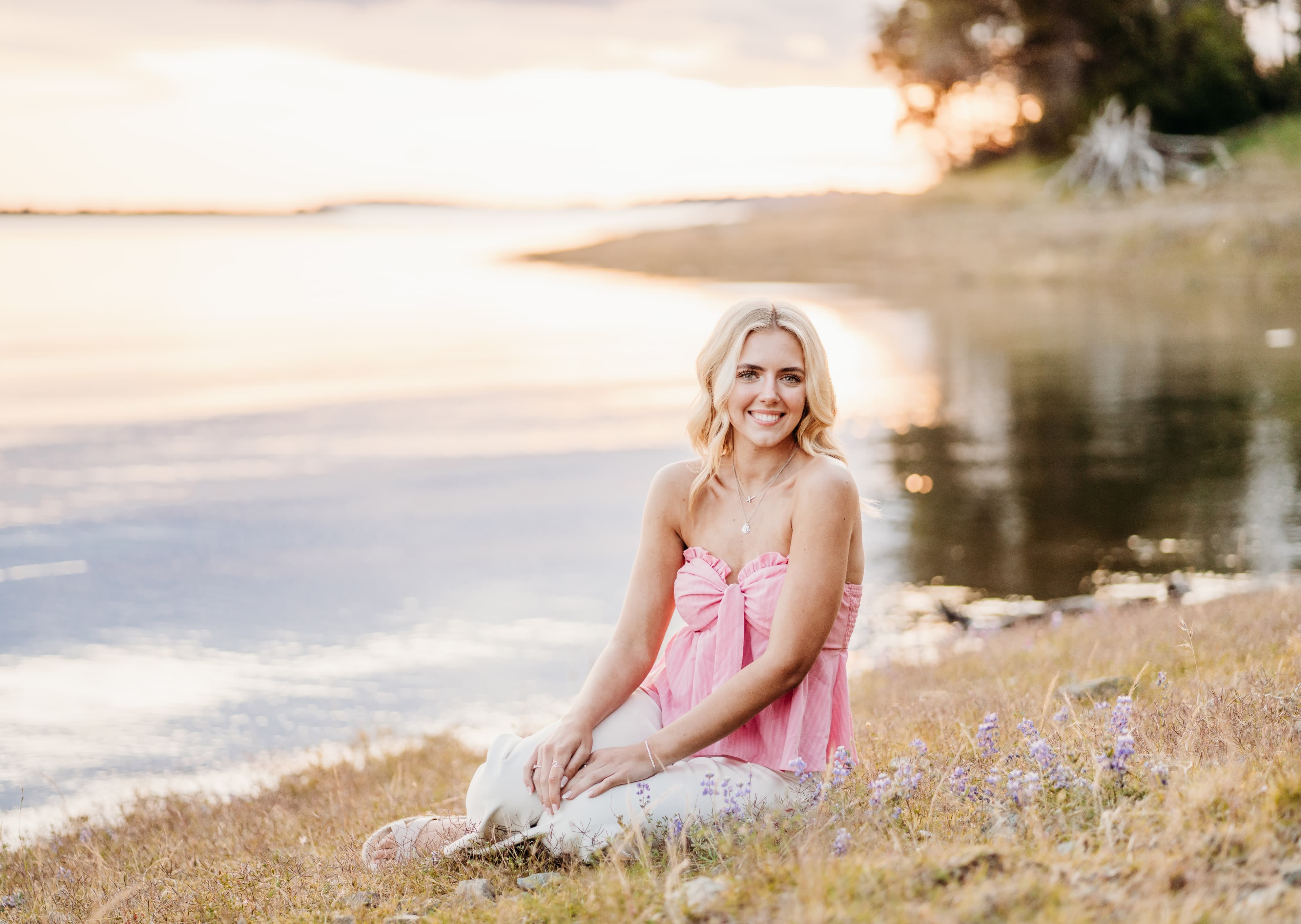 Young woman in a pink top sits on a lakeshore among wildflowers at sunset, smiling