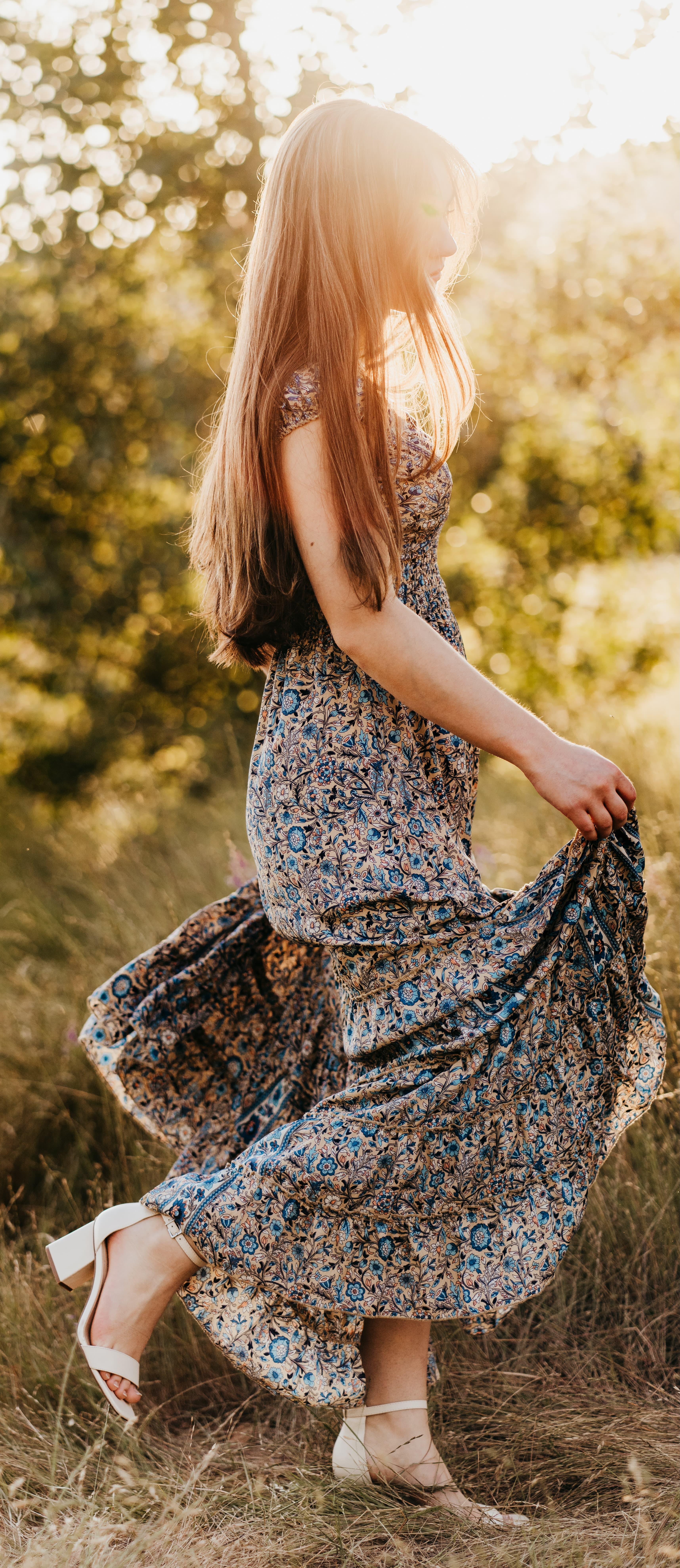 Young woman twirls in a blue floral dress in a sunlit field, one leg kicked up mid-motion