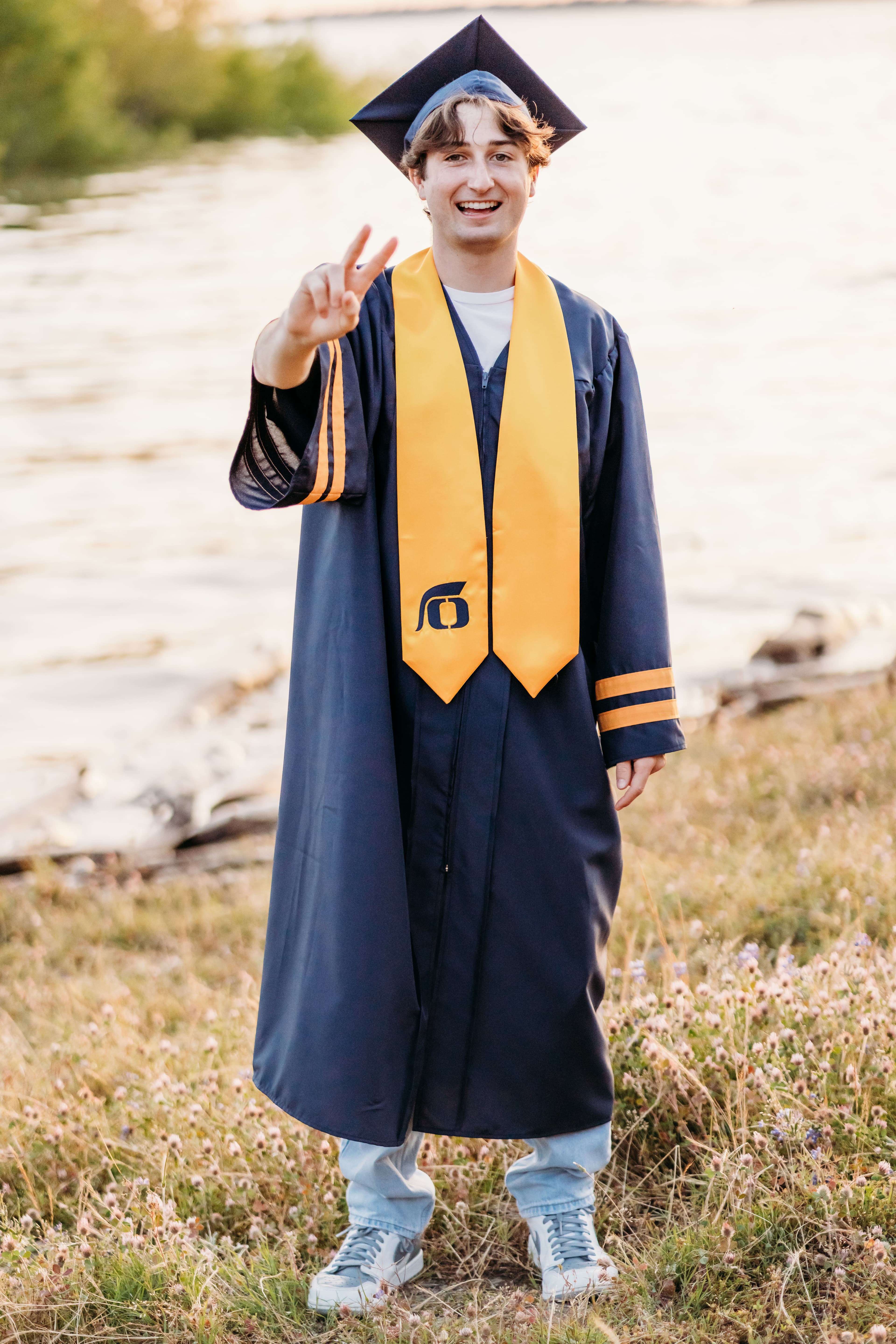 Graduate in cap, gown, and gold stole gives a peace sign by the lake