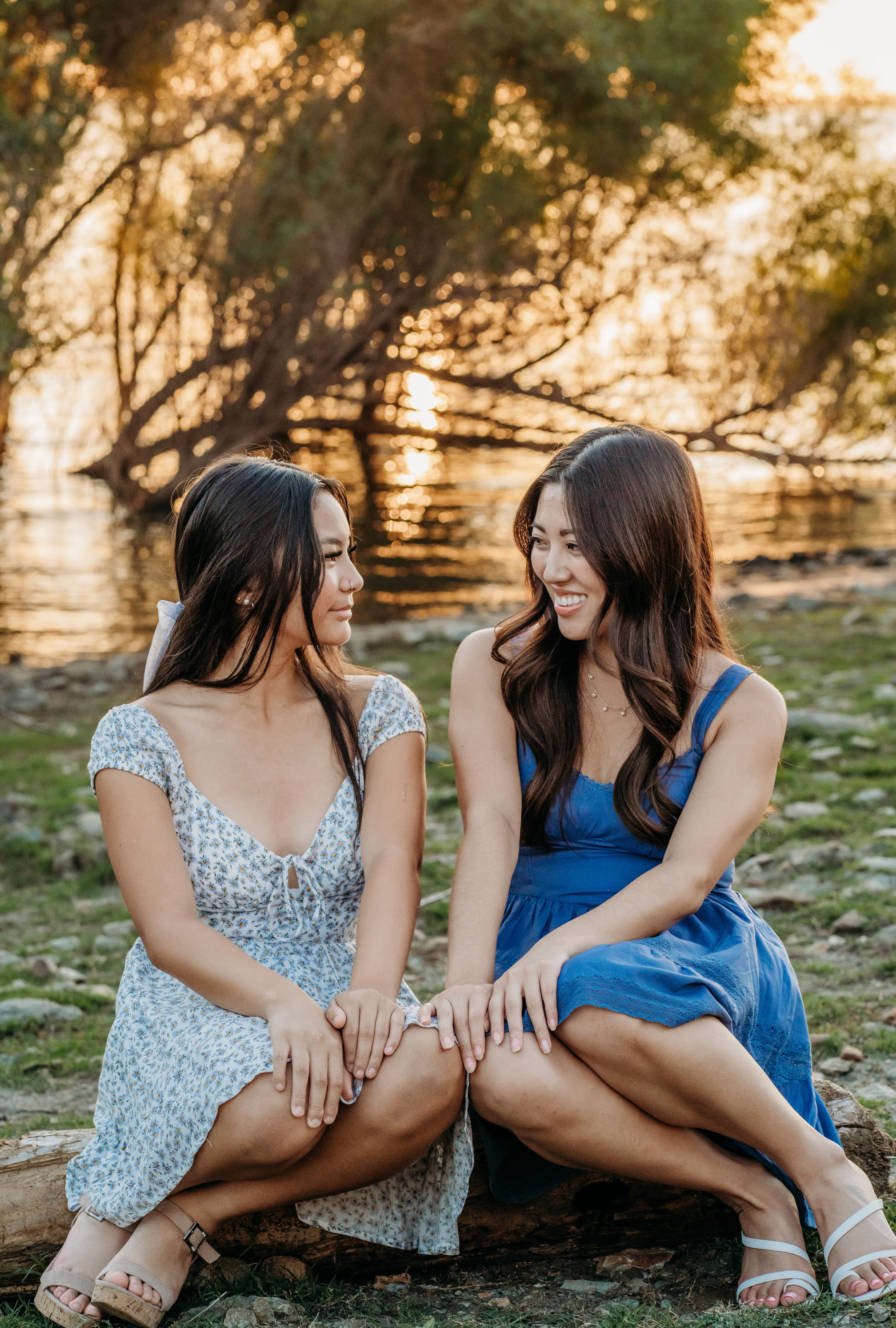 Two sisters sit together on a log by a lake at sunset, hands stacked on their knees, smiling at each other