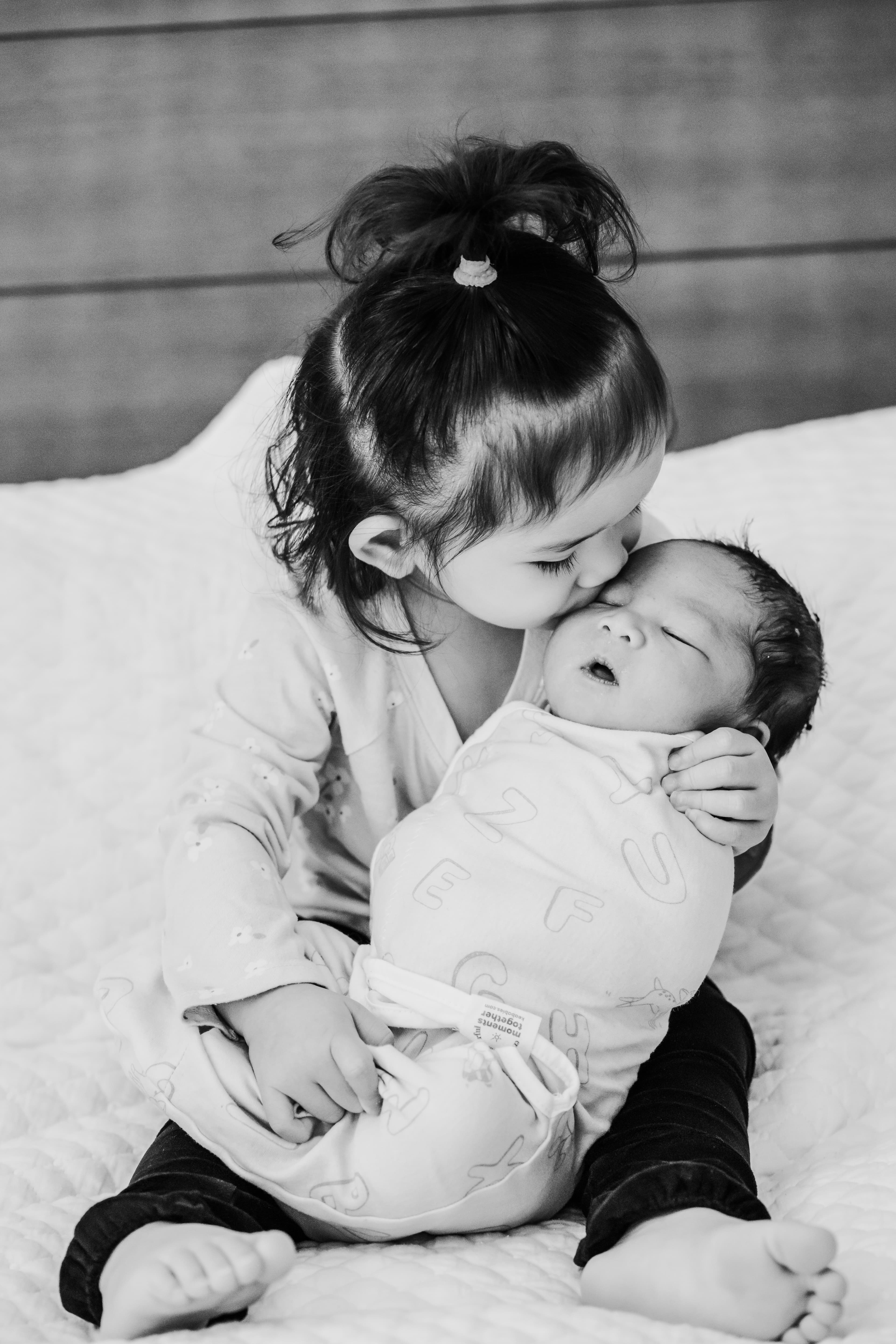 Black-and-white photo of a toddler tenderly kissing her newborn sibling's forehead