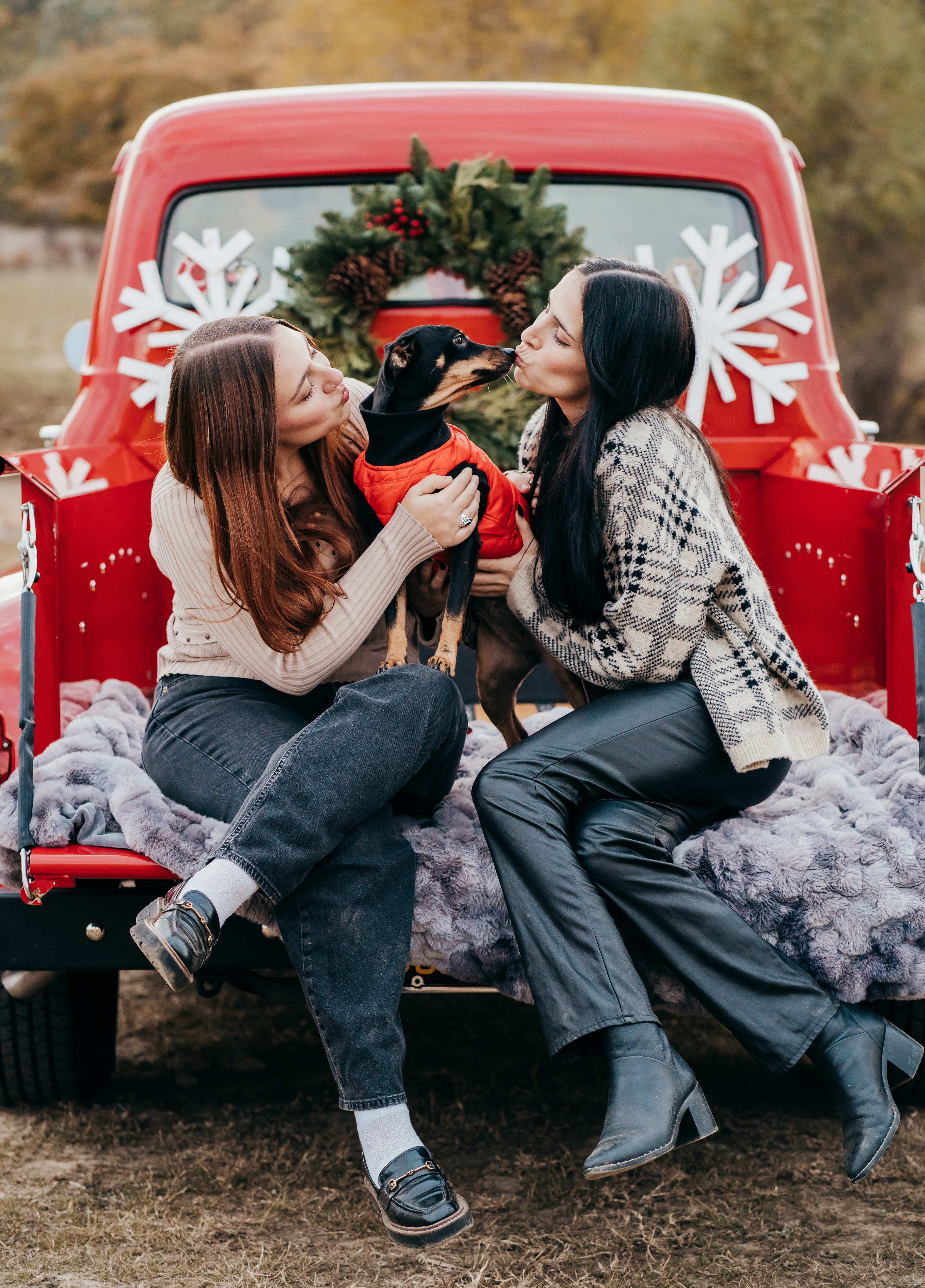 Two women each lean in to kiss a small dog in an orange coat, seated on a red truck tailgate