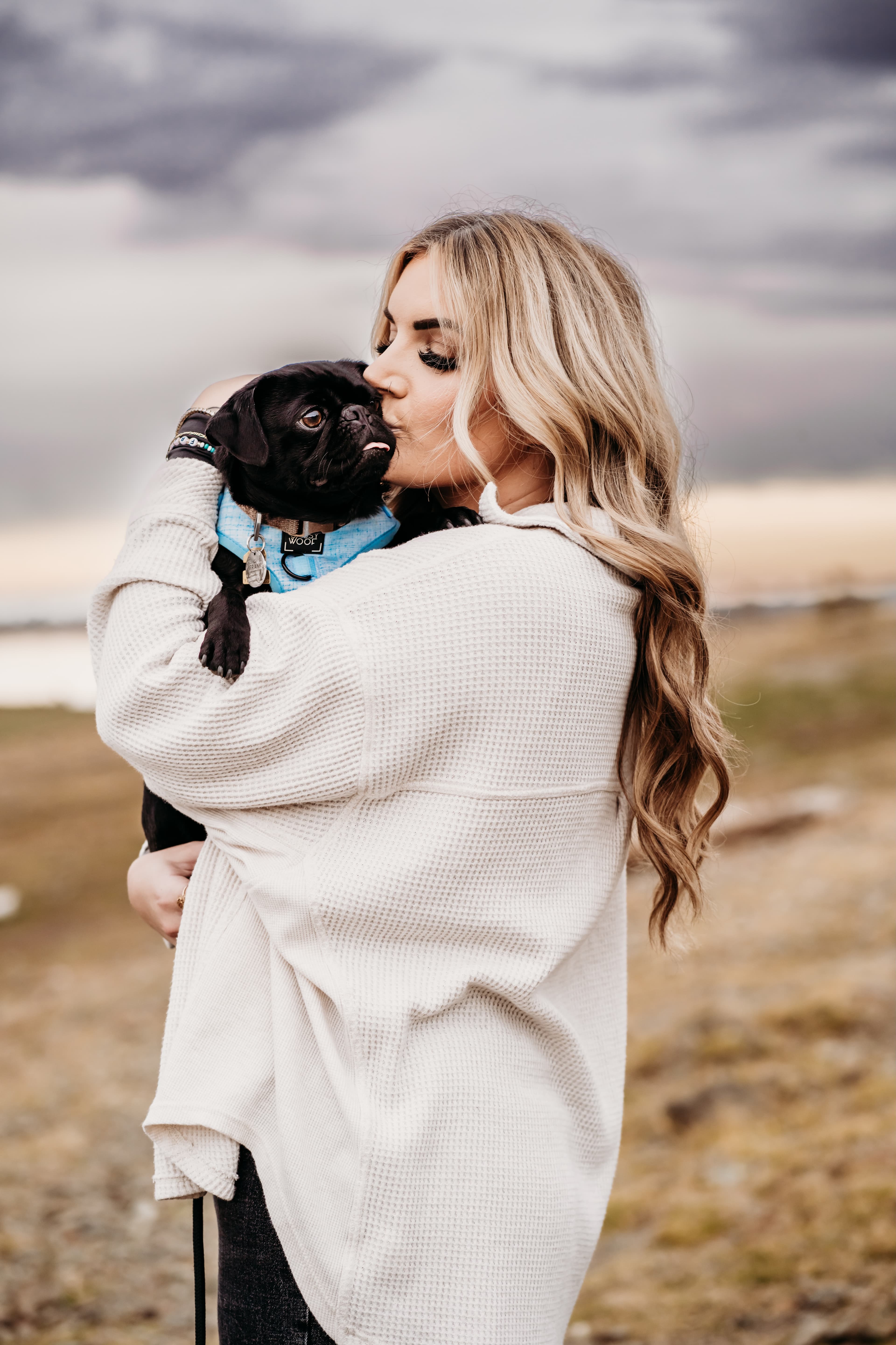 Woman in a cream sweater cradles and kisses a black pug against a moody sky