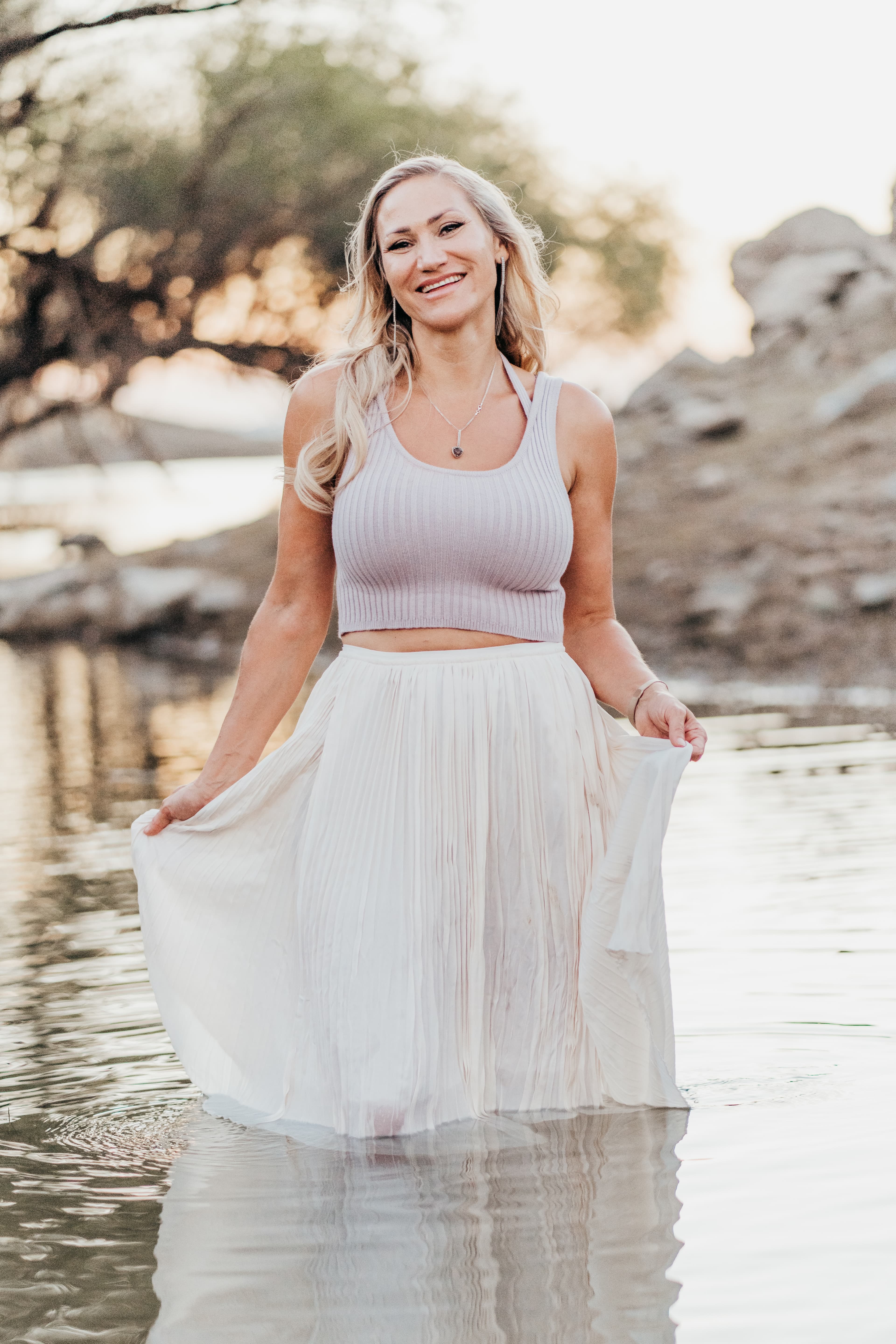 Woman in a flowing white skirt wades in shallow water at golden hour, smiling
